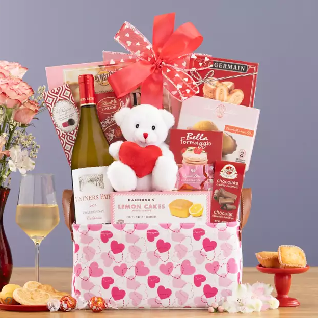 Gift basket with wine, teddy bear, and snacks on a table with a glass of wine.